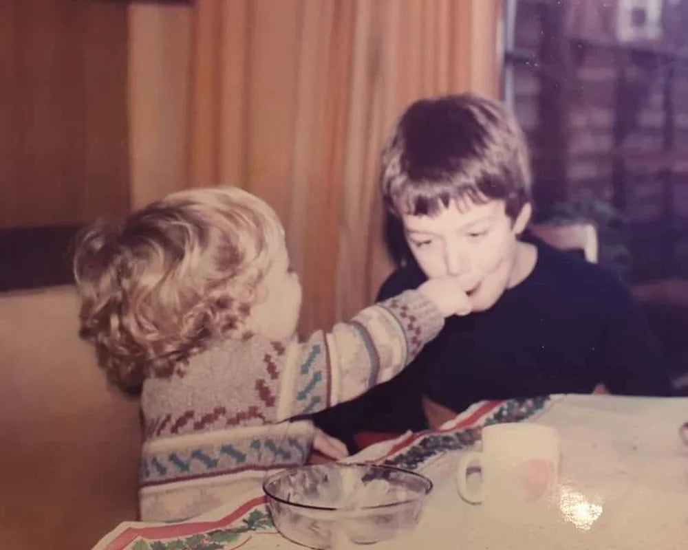 Two kids at the kitchen table. The little one, in a patterned jumper, is feeding the bigger one.